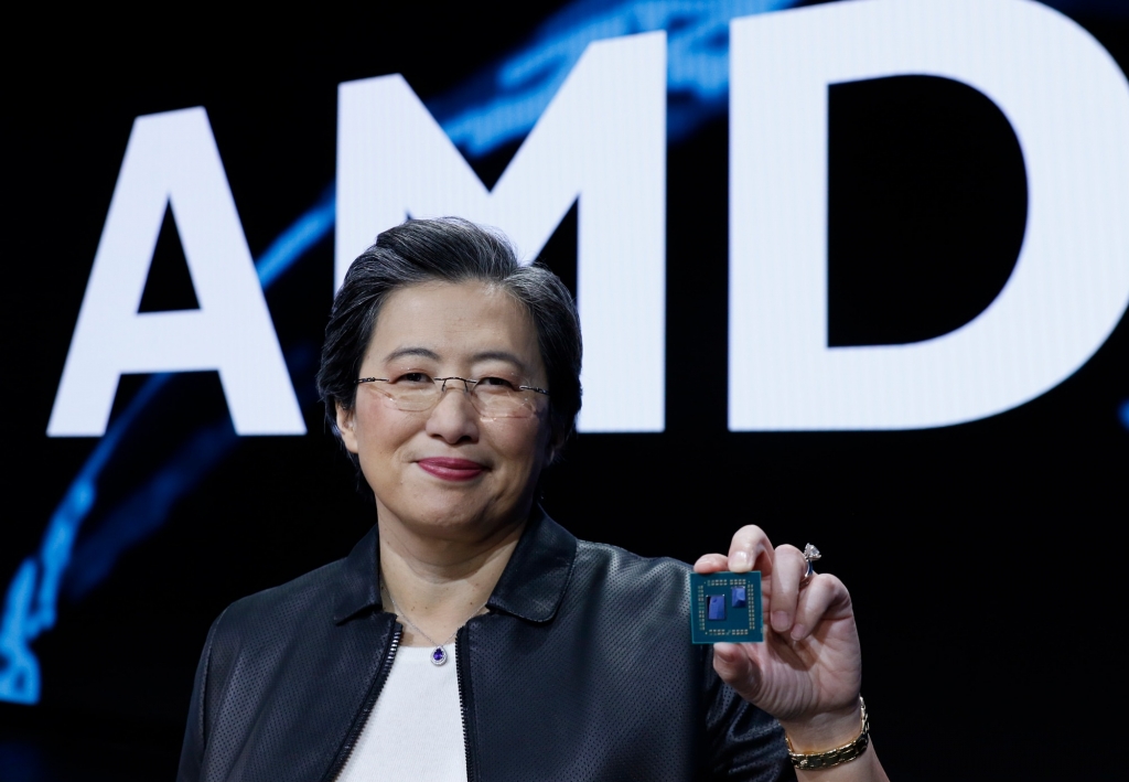 Lisa Su holds a microprocessor created by AMD, with the company letters in the background behind her.