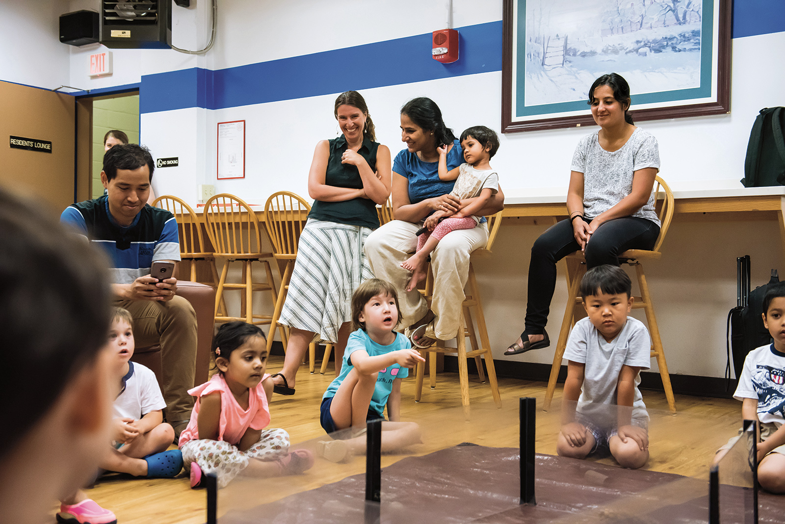 Two women, one holding a small child, look upon a group of small children sitting on the floor